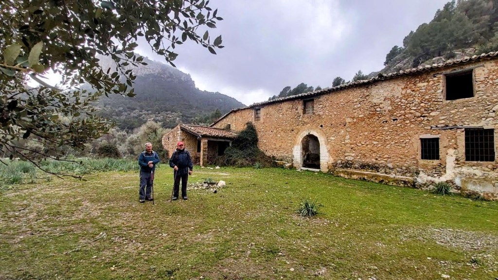 Dos personas de pie frente a las Casas de Olí Clar, antiguas casas de piedra en un área natural con montañas al fondo.