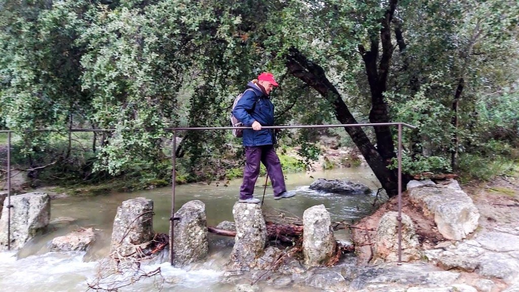 Un senderista cruza el Torrente de Solleric saltando sobre piedras mientras sostiene una barra de metal para equilibrio, rodeado de vegetación.
