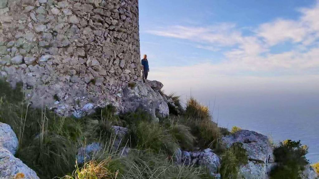 Vista de la Torre de Na Seca con una persona de pie junto a ella, rodeada de rocas y vegetación, con el mar al fondo y un cielo ligeramente nublado.