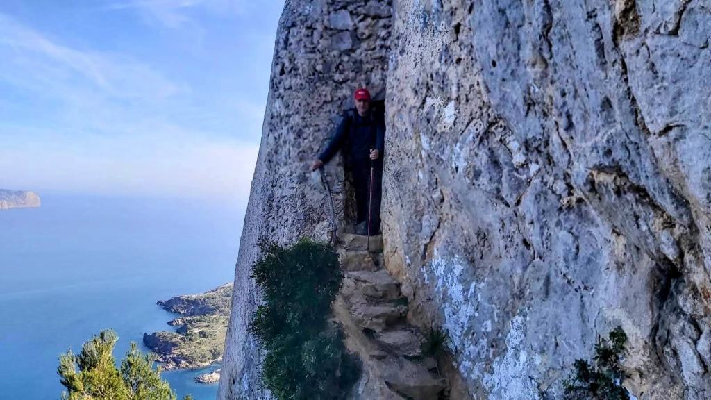Un senderista camina por un estrecho camino de piedra al borde de un acantilado, con vistas al mar y a la costa.