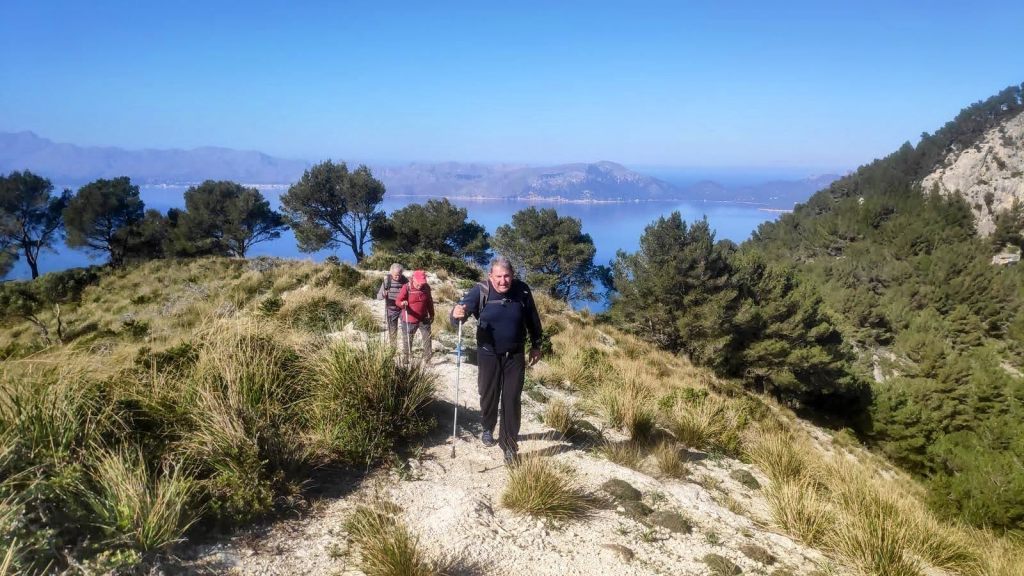 Grupo de senderistas caminando por un sendero montañoso con vista al mar y montañas en el horizonte.