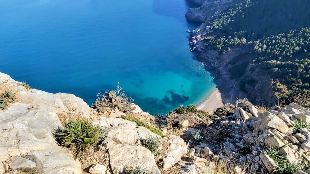 Vista panorámica desde un acantilado hacia la Platja des Coll Baix con aguas cristalinas y rocosas.