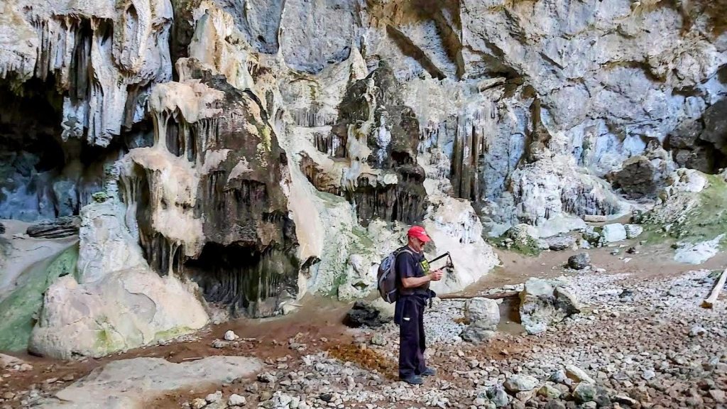 Interior de la Cueva de Migdia, con estalactitas y un senderista con mochila y gorra roja, fotografiando el paisaje rocoso.