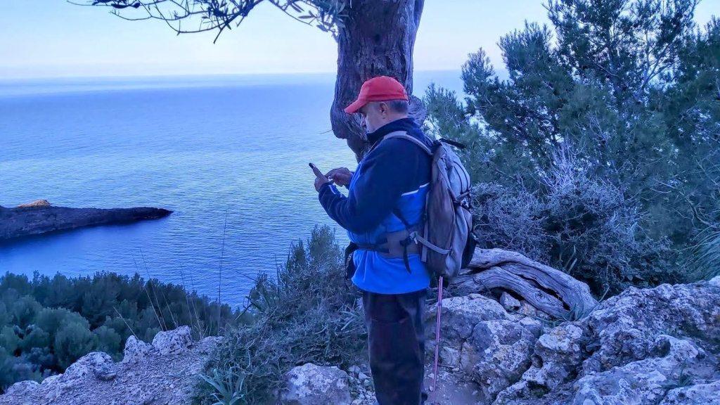 Persona con mochila y bastón de senderismo, usando un teléfono móvil, de pie al borde de un acantilado con vistas al mar y rodeado de árboles en un paisaje montañoso.