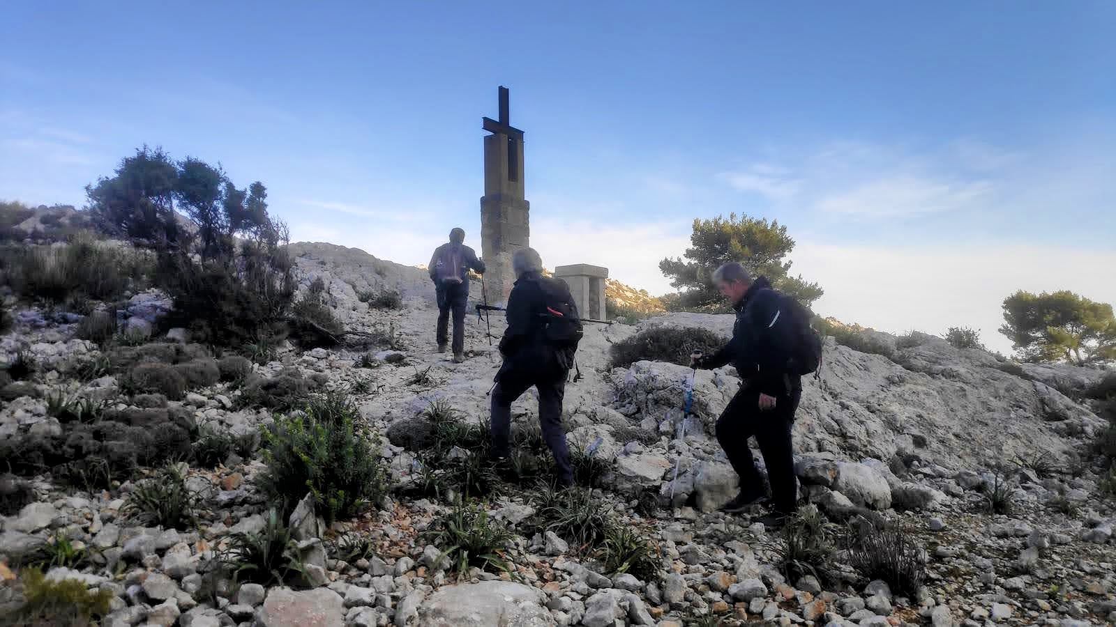 Grupo de senderistas ascendiendo hacia Coll des Cards Colers, con una cruz en el fondo. El paisaje rocoso y la vegetación típica de la zona son visibles bajo un cielo despejado.