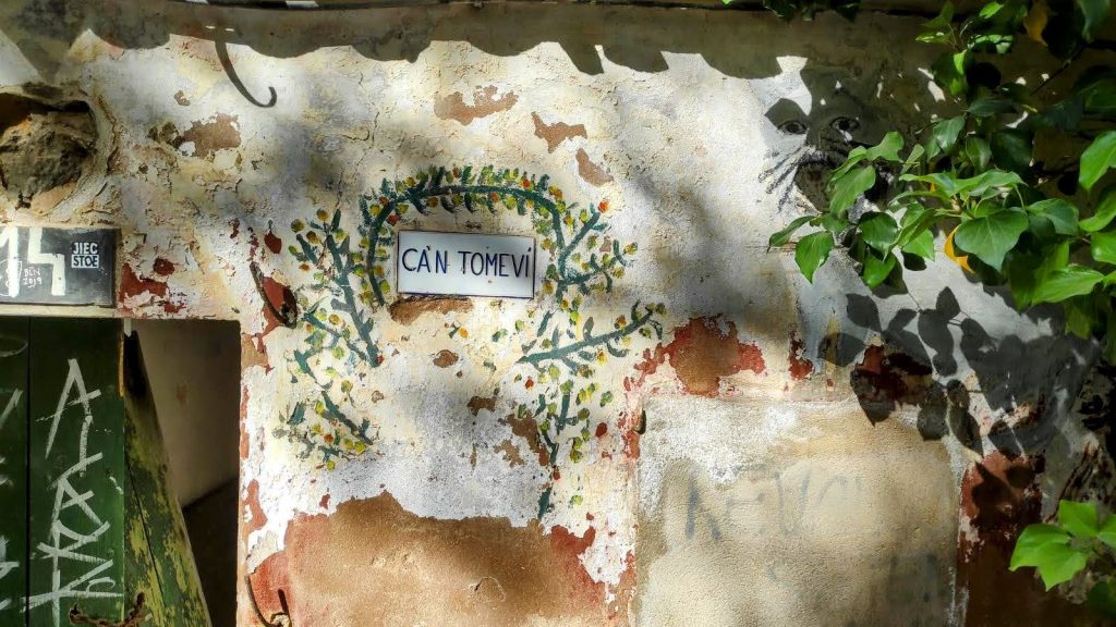 Fachada de la antigua construcción de Can Tomeví con un letrero identificativo y decoraciones naturales en la pared.
