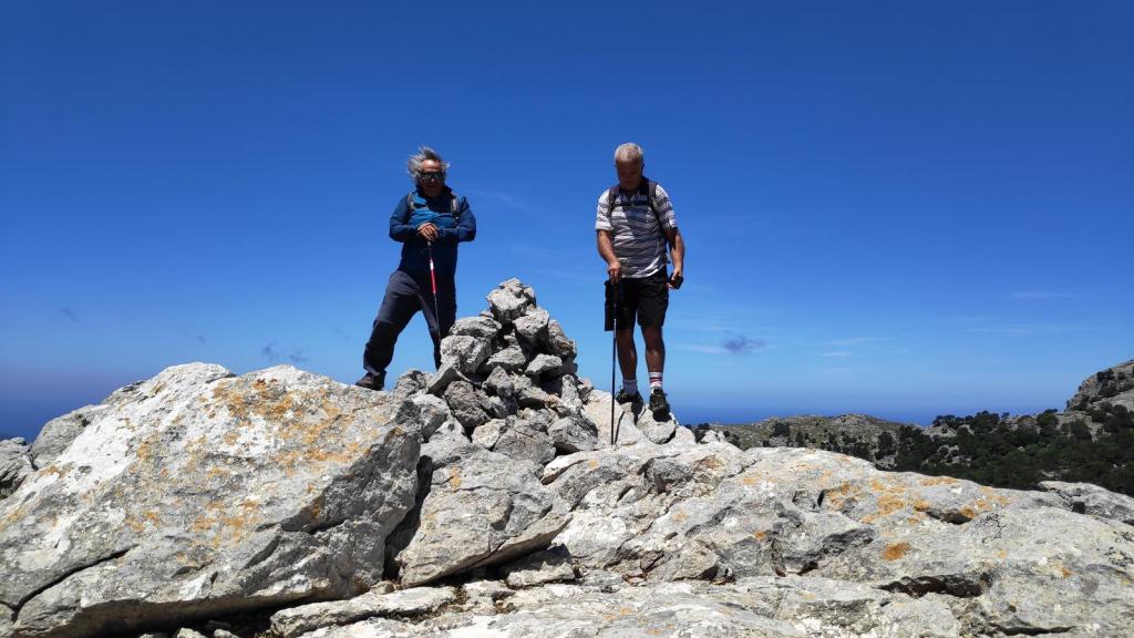 Dos senderistas en la cima del Puig de n’Angelé, rodeados de rocas y un cielo azul claro, disfrutando de las vistas panorámicas.