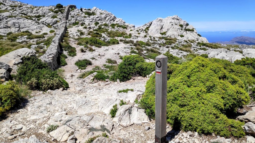 Camino rocoso en la Serra de Tramuntana, Mallorca, con un poste indicativo y vegetación baja entre formaciones rocosas.