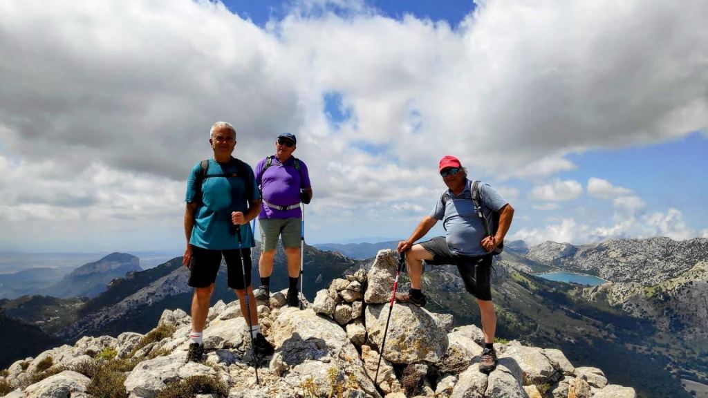 Grupo de tres excursionistas en la cima de una montaña con un paisaje montañoso y un embalse al fondo, en un día nublado.