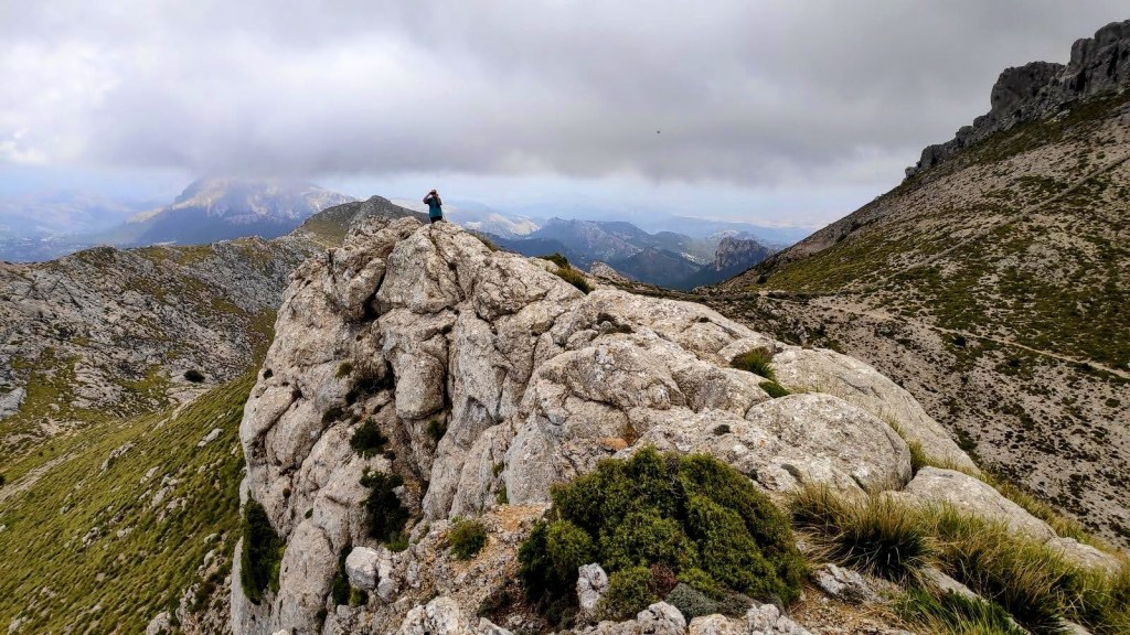 Una persona de pie sobre una formación rocosa en la cima de una montaña, rodeada de un paisaje montañoso con nubes en el cielo.