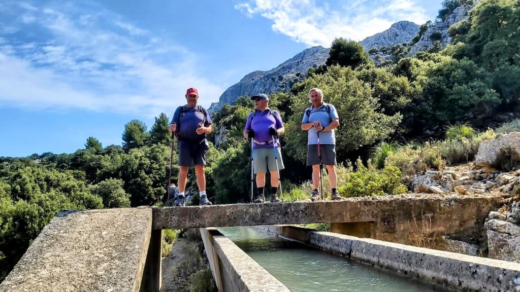 Tres excursionistas de pie sobre un puente de hormigón que cruza un canal de agua, con un paisaje montañoso y vegetación densa al fondo.