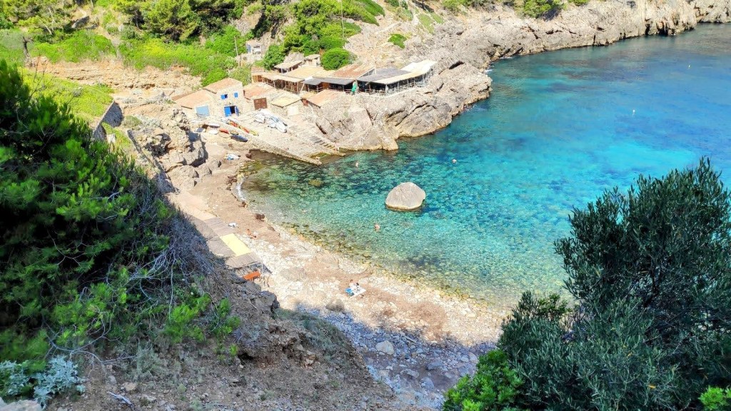 Vista panorámica de la Cala de Deiá, con aguas transparentes y formaciones rocosas, rodeada de vegetación, mostrando un pequeño embarcadero y cabañas.