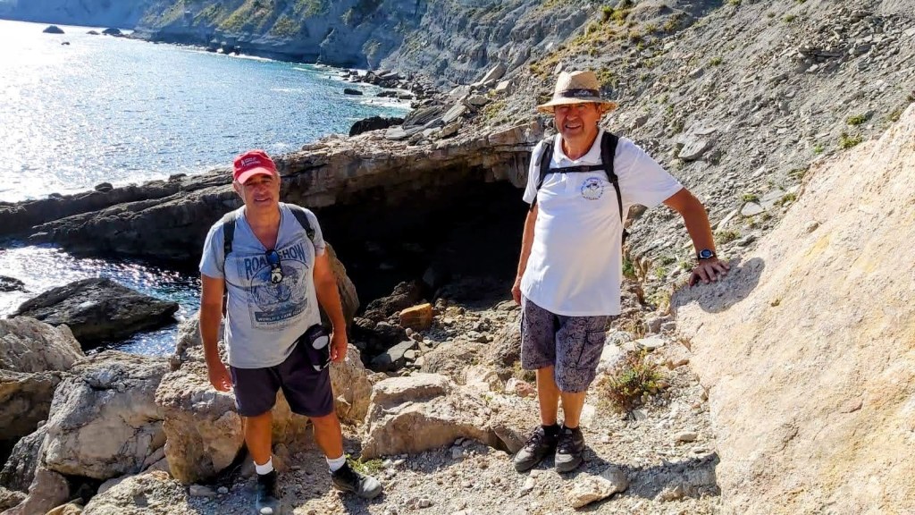 Dos excursionistas en una zona rocosa cerca del mar, junto a la entrada de la Cova de Na Bernarda.