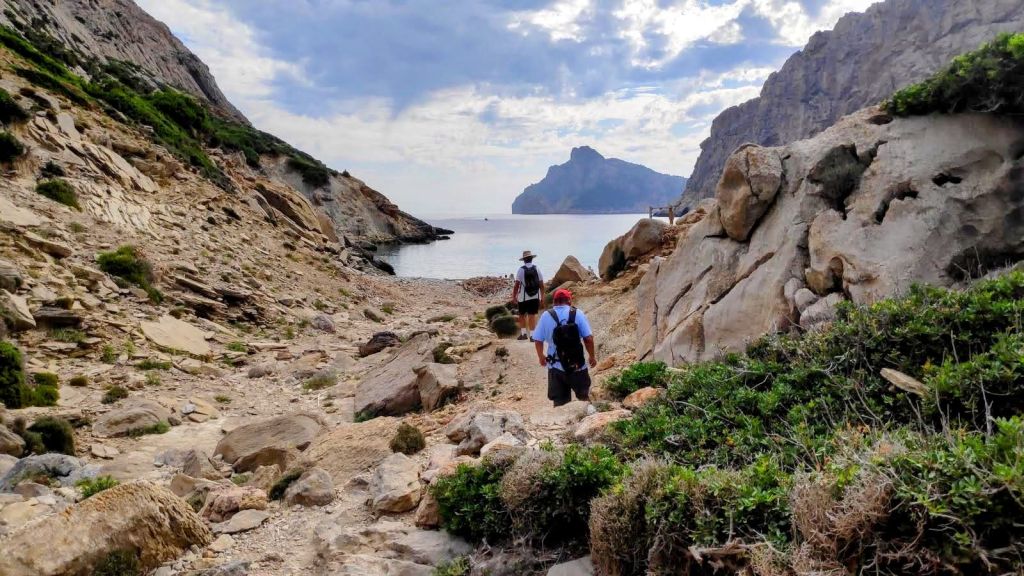 Sendero que conduce a la playa de Cala Bóquer, con montañas y un grupo de personas caminando por el camino rocoso.