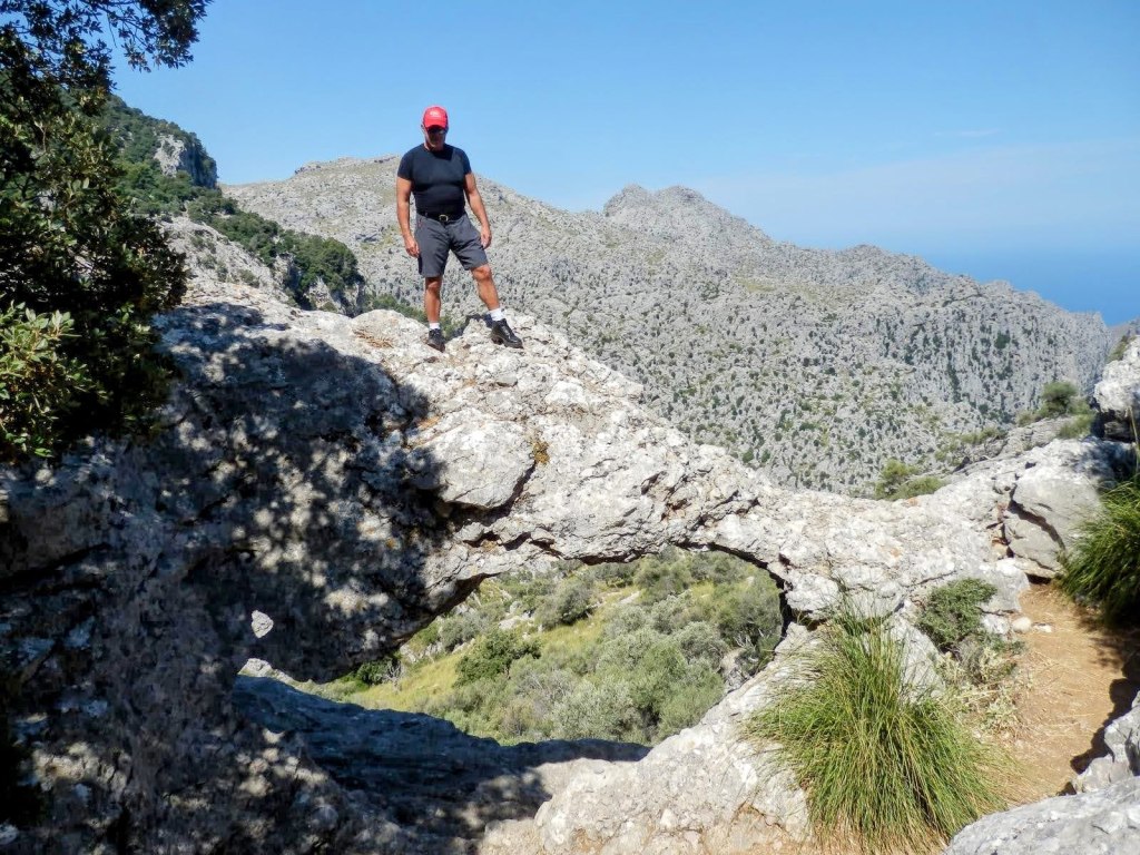 Hombre de pie sobre una roca con forma de arco, en un paisaje montañoso de la Serra de la Tramuntana, con el cielo despejado al fondo.