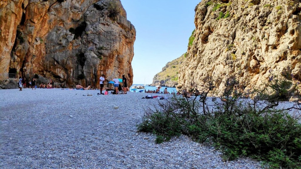 Playa en Sa Calobra, rodeada de altas formaciones rocosas y gente disfrutando del sol y el mar.
