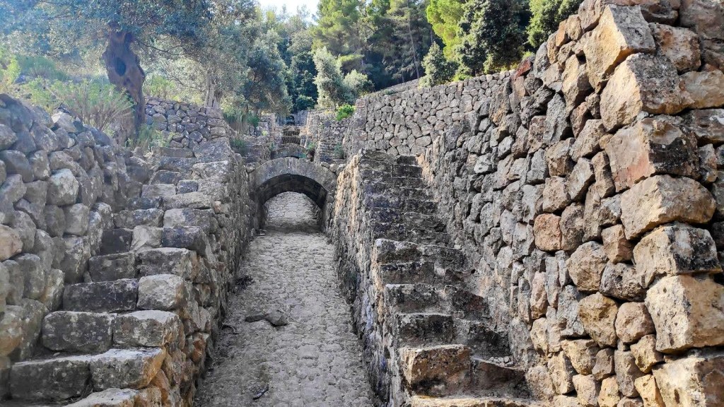 Sendero empedrado con escalones de piedra y un arco de piedra en un entorno natural con vegetación.