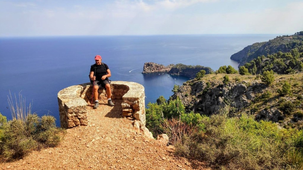 Ruta Sa Foradada-Miramar. Caminando por Mallorca Hombre sentado en el Mirador de Sa Ferradura con vista al mar y acantilados, rodeado de vegetación.
