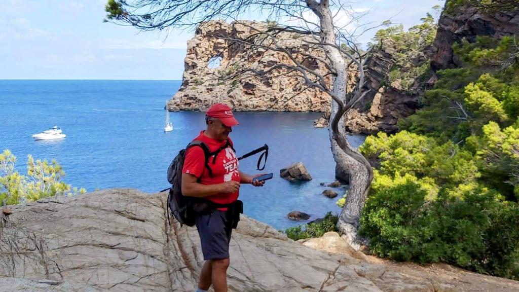 Un senderista observa el paisaje desde el Mirador natural del Camí de la Mar en la Ruta Sa Foradada, con vistas a una formación rocosa en el mar y un barco en la distancia.