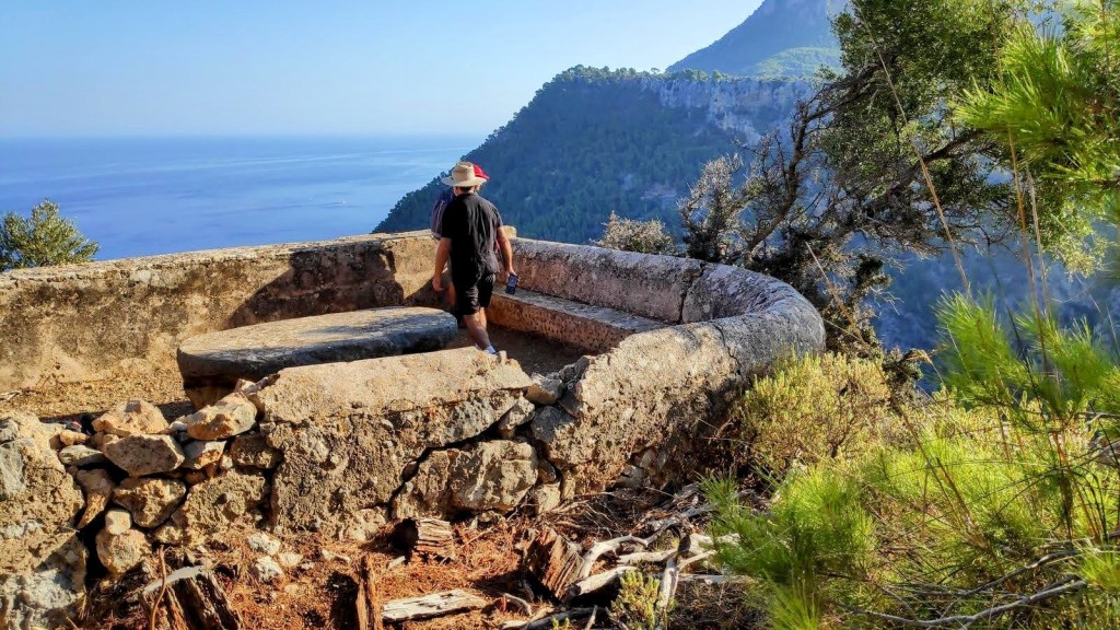 Vista panorámica desde el Mirador des Pi, con un hombre de espaldas de pie sobre una plataforma de piedra. El fondo muestra un mar azul y montañas verdes.