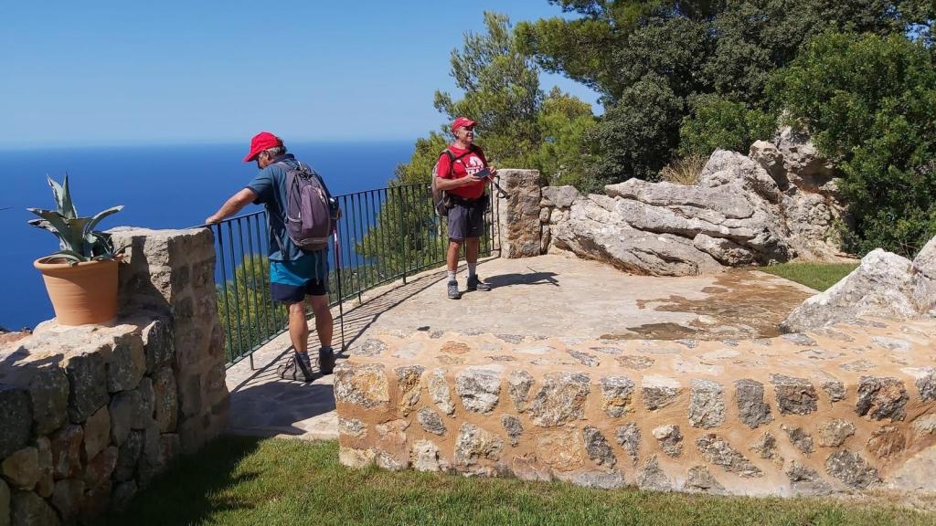 Dos excursionistas con gorras rojas en un mirador con vistas al mar, rodeados de vegetación y rocas, disfrutando del paisaje.