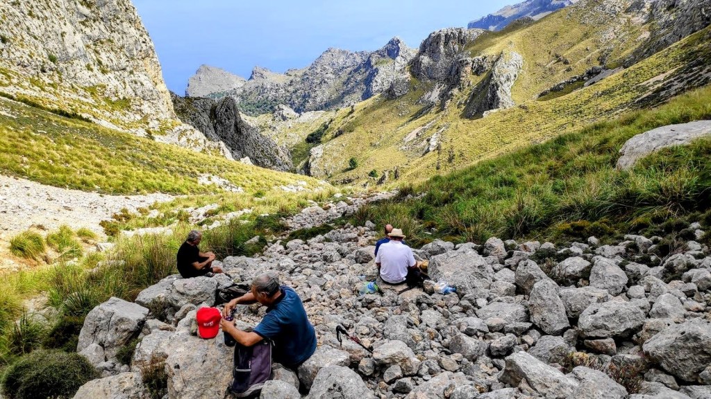 Tres personas descansando entre rocas en la Coma Fosca del Puig Major , con vistas a un valle montañoso y un cielo despejado.