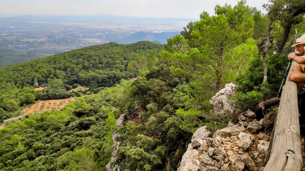 Vista panorámica desde un mirador en la montaña, con un hombre sentado en una barandilla de madera. Se observa un paisaje de bosque denso con árboles y un valle al fondo, donde se aprecian áreas cultivadas y una ciudad distante.