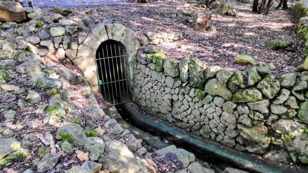 Fuente de Llinars, una estructura de piedra con una entrada con reja, rodeada de hoja y rocas en un entorno natural