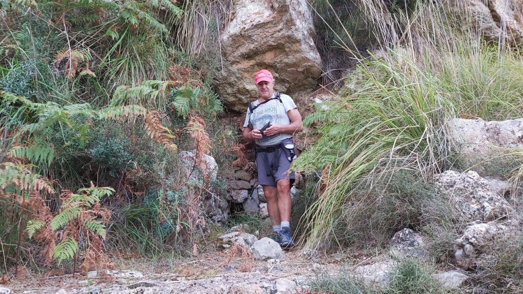 Hombre de pie  delante de la Font Subauma, entre vegetación en una ruta montañosa, sosteniendo un objeto, vestido con ropa de senderismo.