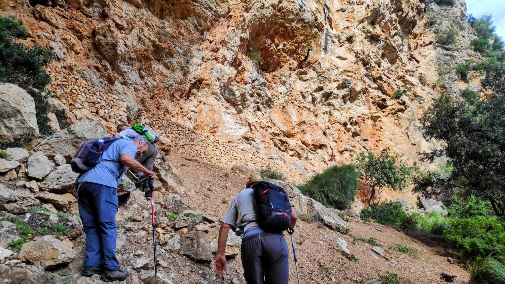 Dos excursionistas subiendo por un terreno rocoso a la balma Na Mamelluda, rodeados de naturaleza y paredes de roca.