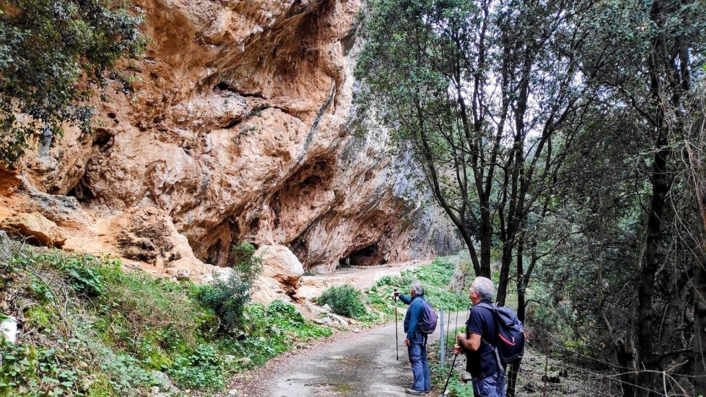 Dos senderistas observan una cueva grande y rocosa en un sendero rodeado de vegetación en la Serra de Tramuntana.