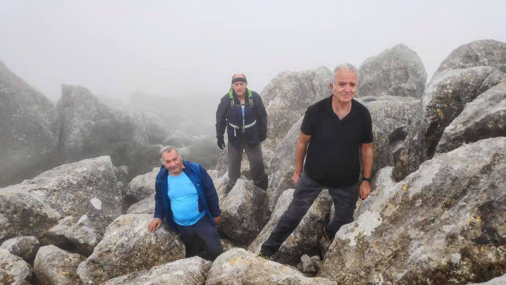 Ruta del Puig de n'Alí-Caminando por Mallorca Grupo de tres excursionistas en la cima del Puig de n'Alí, rodeados de grandes rocas y niebla.