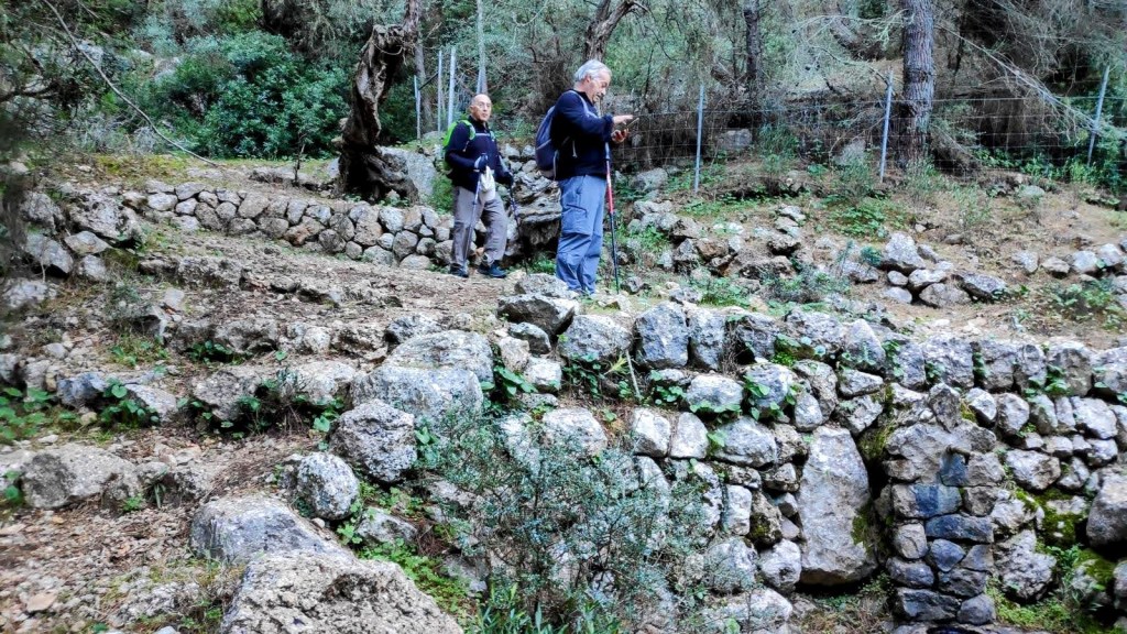 Dos excursionistas en un paisaje montañoso, rodeados de muros de piedra y vegetación, en la Font de sa Senyora