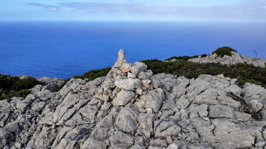 Ruta Cap de Catalunya-Pal de Formentor-Caminando por Mallorca Cima de la Penya de la Cova dels Morts con un hito de piedras en un paisaje rocoso, con vista al mar azul en el fondo.