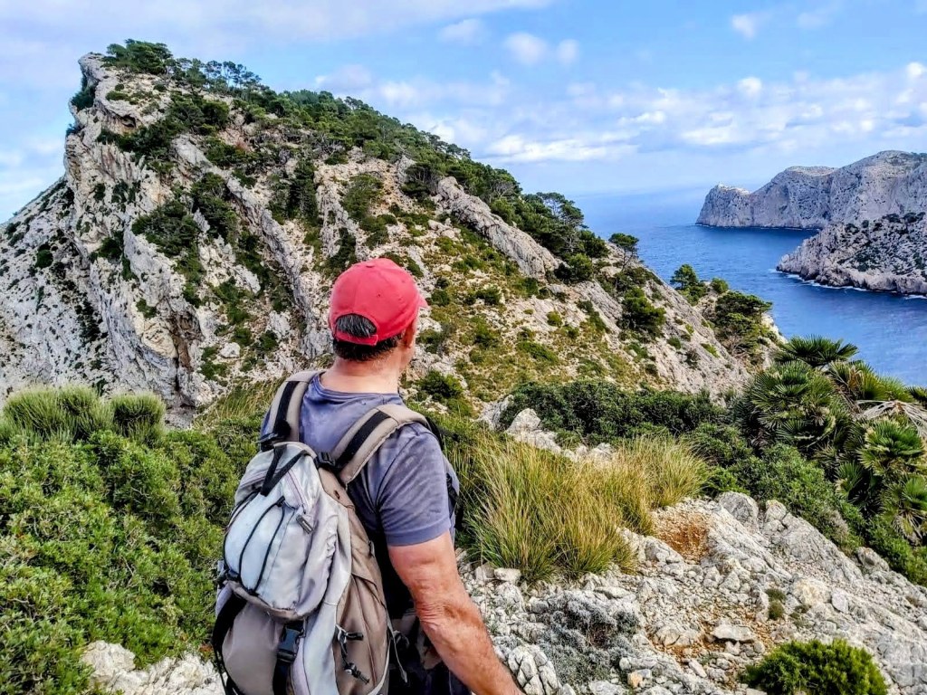 Vista desde una montaña que muestra un sendero rocoso y una costa impresionante, con un hombre que observa el paisaje, usando una mochila y una gorra roja.