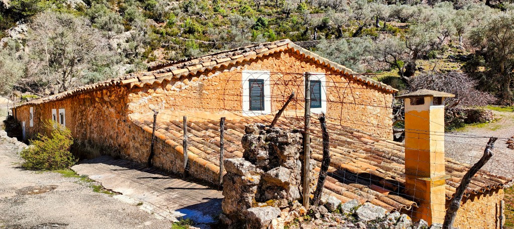 Casa de campo de estilo tradicional, con paredes de piedra y un tejado de tejas, rodeada de vegetación y un paisaje montañoso.
