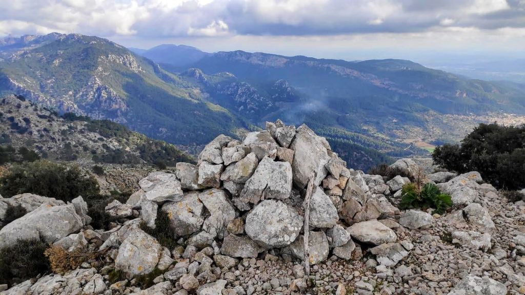 Pila de piedras en la cima del Puig de Sa Font, con un paisaje montañoso y nubes en el cielo de fondo.