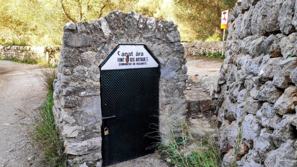 Entrada de la Font de ses Artigues, con una puerta de metal y una estructura de piedra, rodeada de vegetación y un camino de tierra.