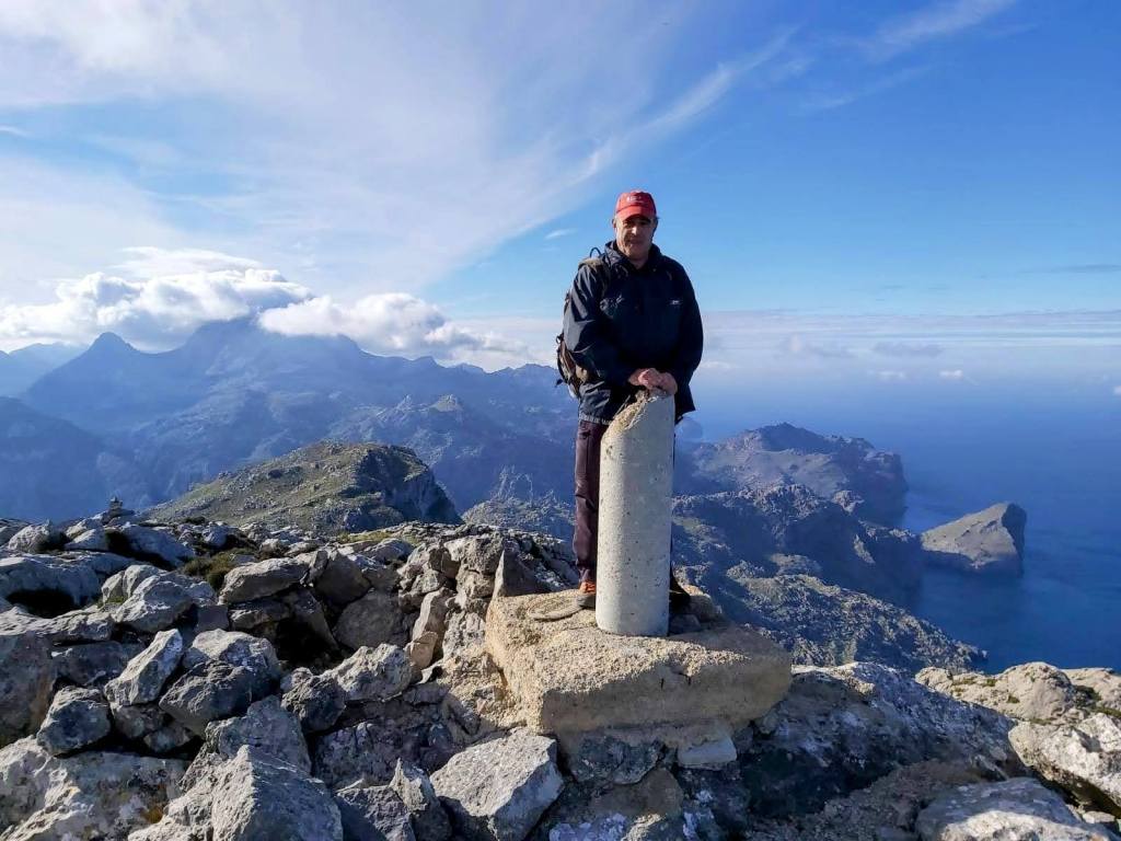Persona de pie en la cima del Puig Roig, rodeada de montañas y vistas panorámicas del paisaje de Mallorca, con un marcador de piedra a su lado.