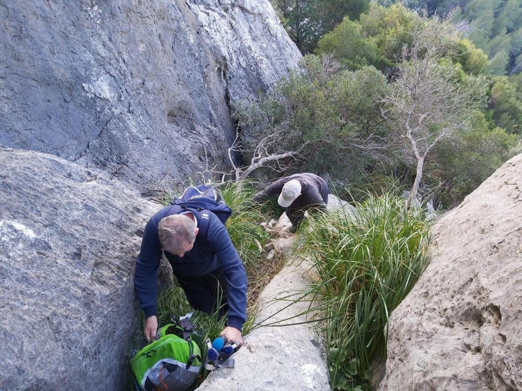 Dos personas explorando un paso rocoso en un entorno montañoso, rodeadas de vegetación y grandes piedras.