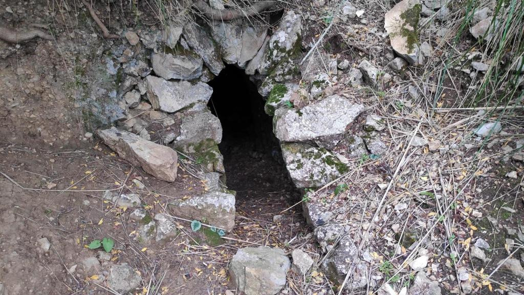 Entrada de una cueva o fuente en la montaña, rodeada de piedras y vegetación.
