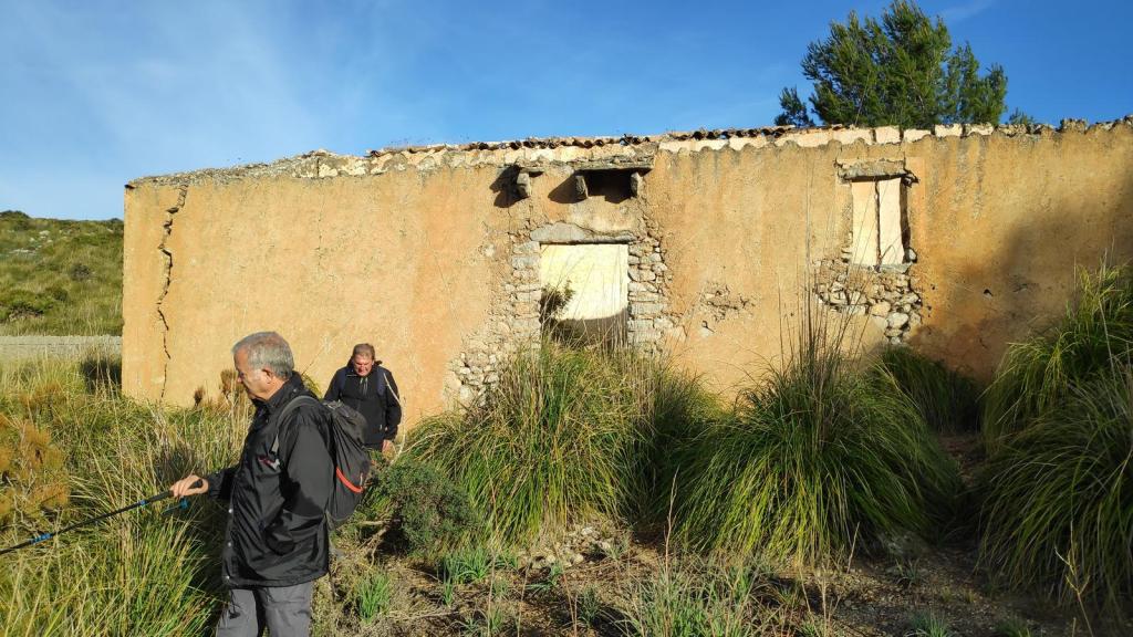 Dos personas caminando cerca de una construcción abandonada, parcialmente derruida, cubierta de vegetación en un entorno montañoso.