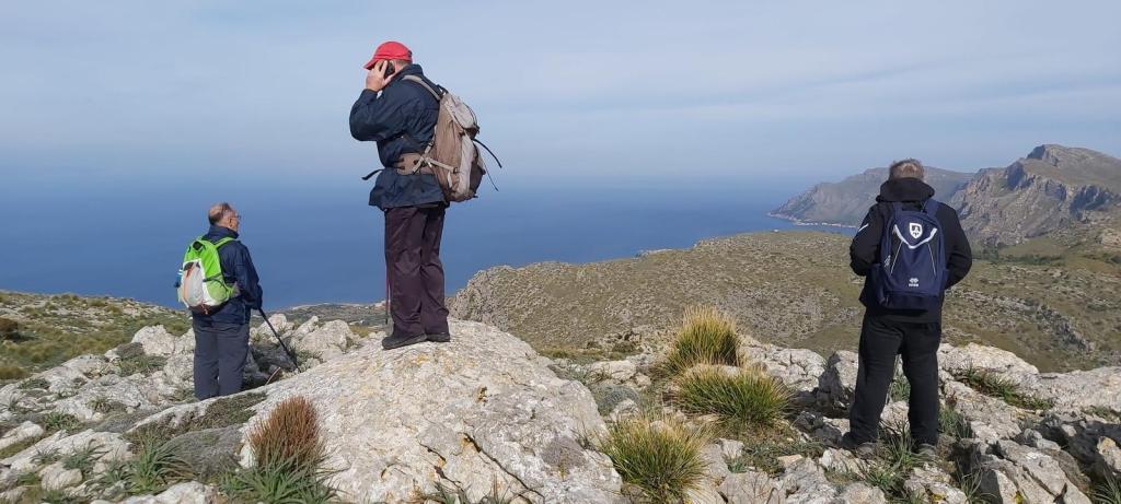 Grupo de excursionistas en la cima del Puig d’en Pelat, con vistas panorámicas al mar y montañas. Uno de ellos está utilizando un teléfono móvil, mientras los otros dos observan el paisaje.