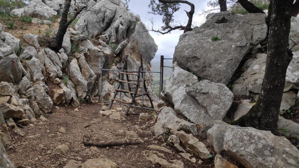 Un sendero montañoso estrecho entre grandes rocas, con una barrera de madera y un cercado metálico al fondo, rodeado de vegetación y cielo nublado.