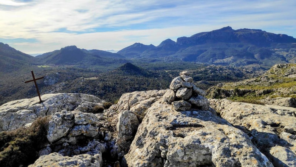 Vista panorámica desde la cima del Puig Rodó, con una cruz de hierro y un hito de piedras en primer plano, rodeado de montañas y un paisaje natural en la Serra de Tramuntana.
