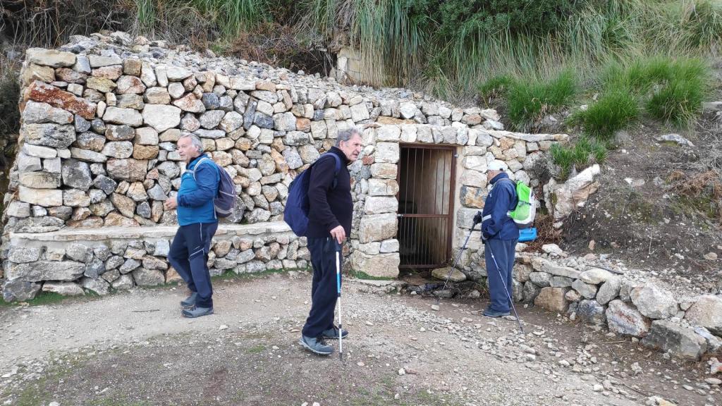 Tres excursionistas junto a la Font d'en Quelota. La estructura parece ser una pequeña edificación rural con una puerta de reja, rodeada de césped y vegetación.