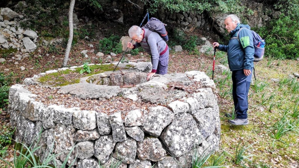 Dos personas observan un pozo de piedra redondeado en un entorno natural, rodeados de vegetación.
