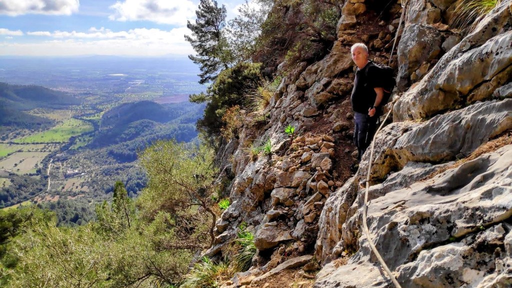 Hombre escalando un paso rocoso empinado con una cuerda de seguridad, rodeado de vegetación y vistas de un valle en el fondo.
