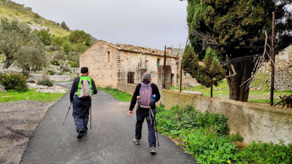 Dos senderistas caminando por un camino asfaltado en una zona rural, con las Casas de Miner Petit y vegetación alrededor.