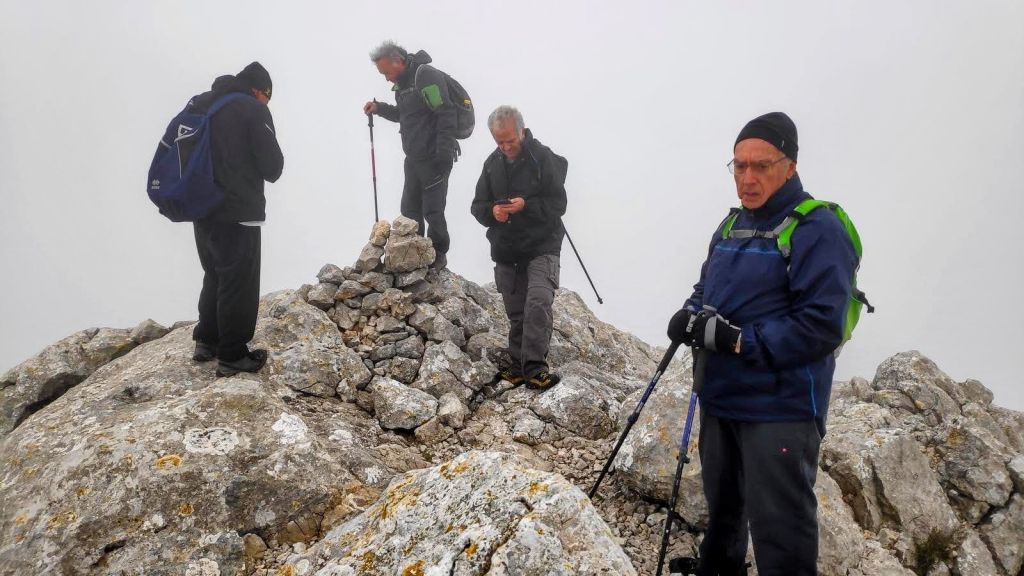 Un grupo de cuatro excursionistas en la cima del Puig de Ca Miner, rodeados de niebla, con un hito de piedras visible en el centro.